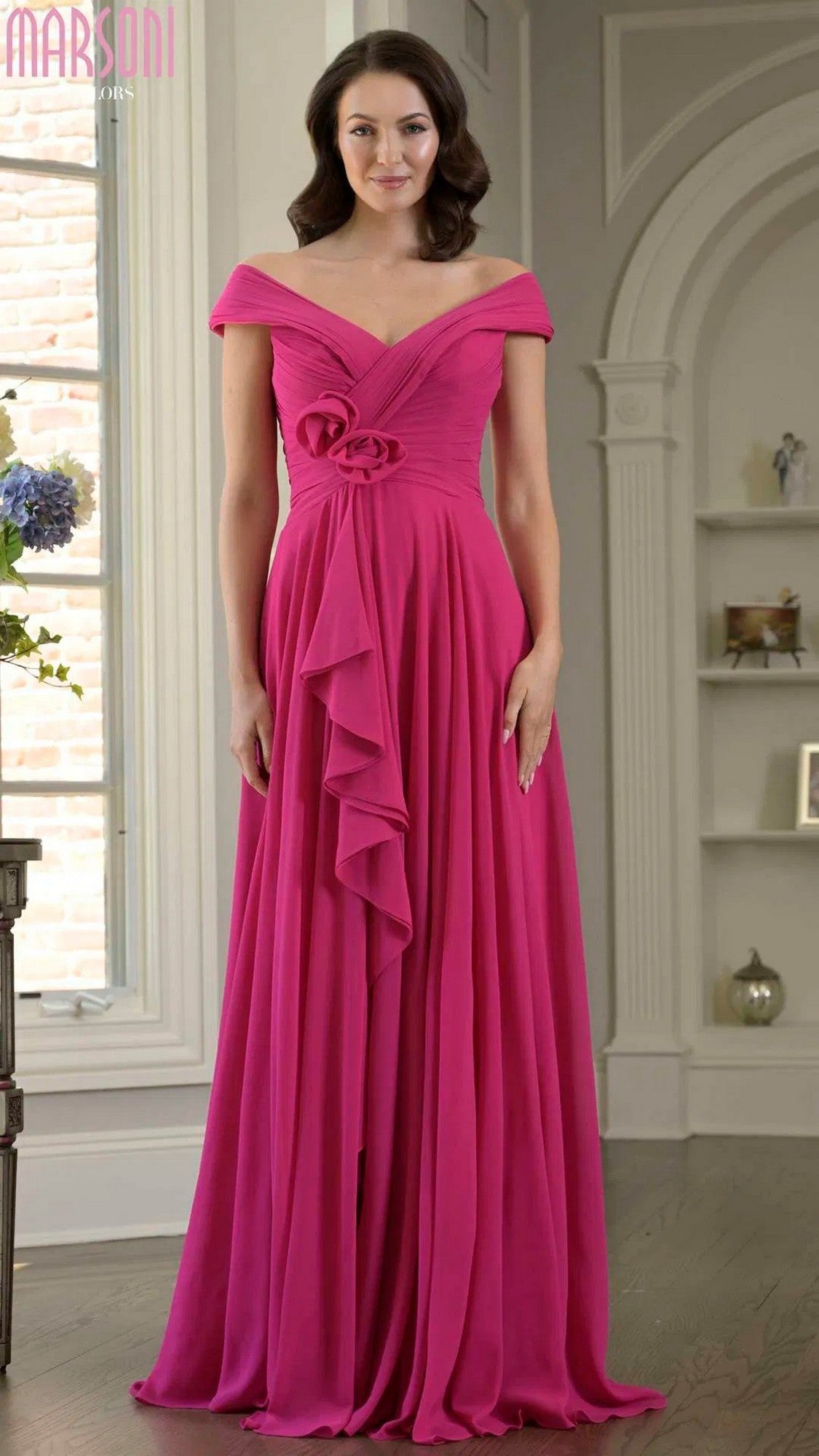 A woman stands indoors wearing the Marsoni M333, a long formal fuchsia dress with floral accents, chiffon fabric, and an off-the-shoulder design featuring a draped front ruffle.